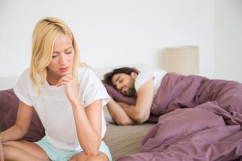 Depressed young woman sitting on the edge of the bed thinking about relationship problems while her partner is sleeping unaware of her sadness.